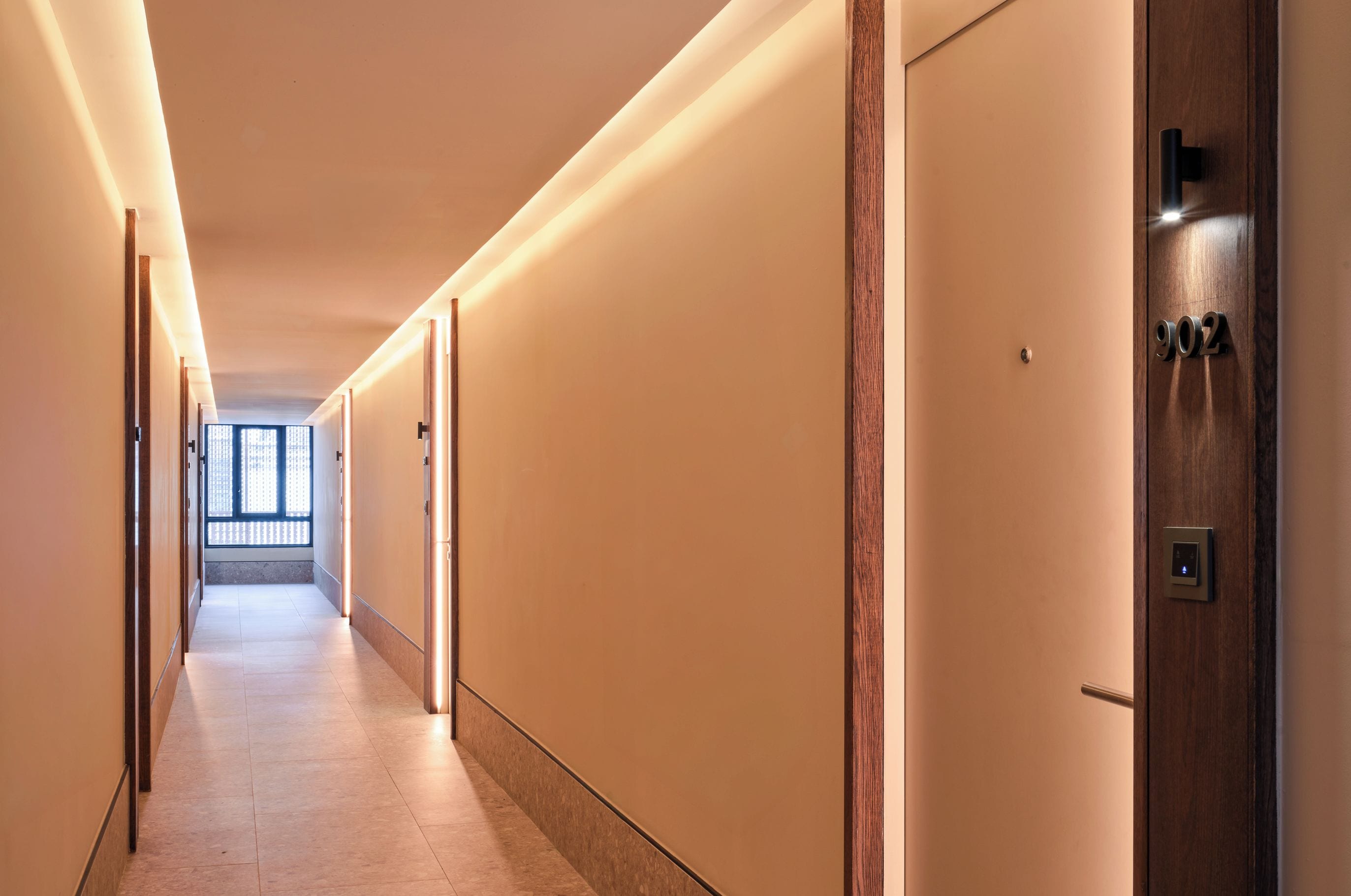 Un long couloir étroit, typique des hôtels, avec des portes d’un côté et une fenêtre au bout.