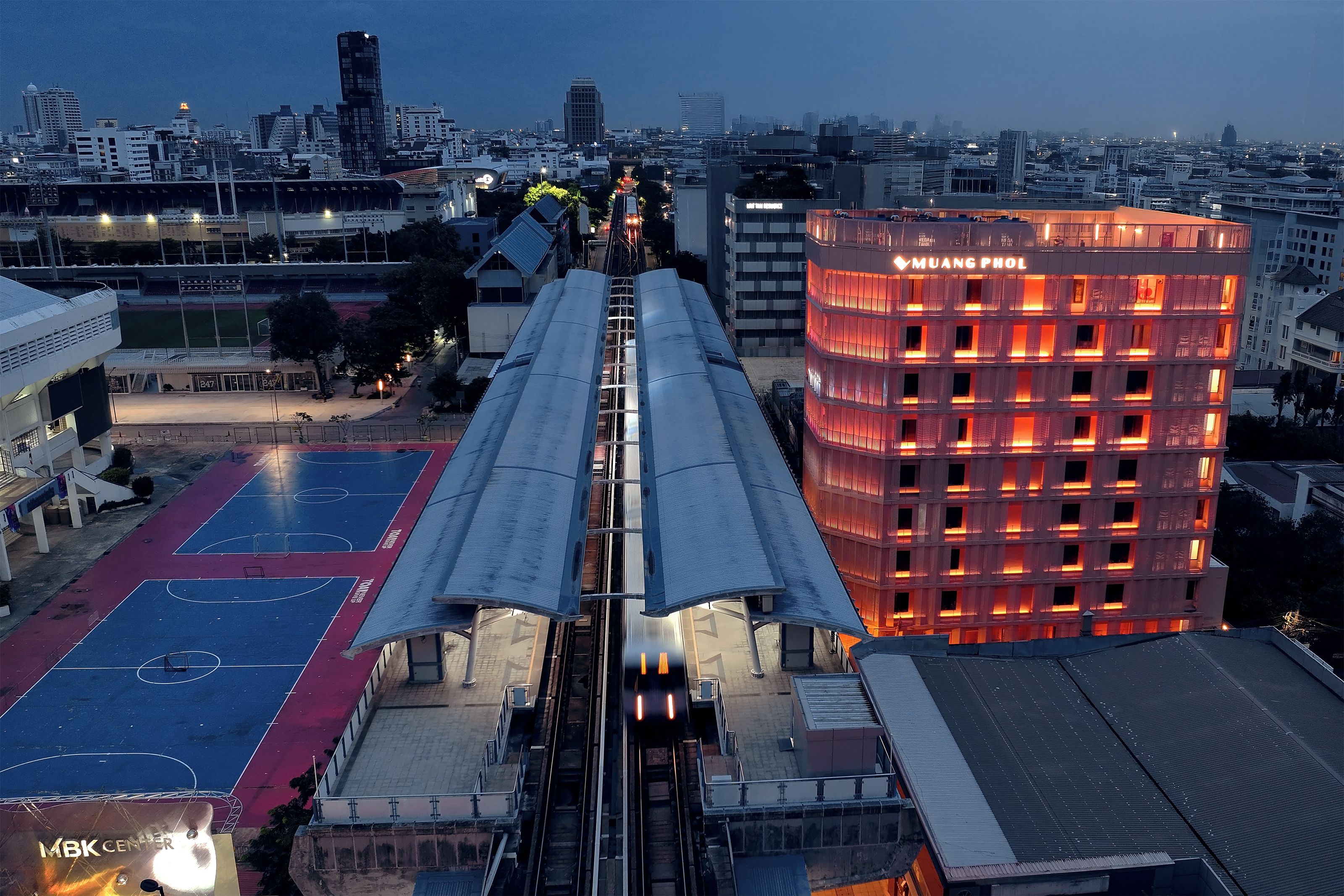 A multi-level parking structure with a train track running through it, surrounded by buildings and a sports court.