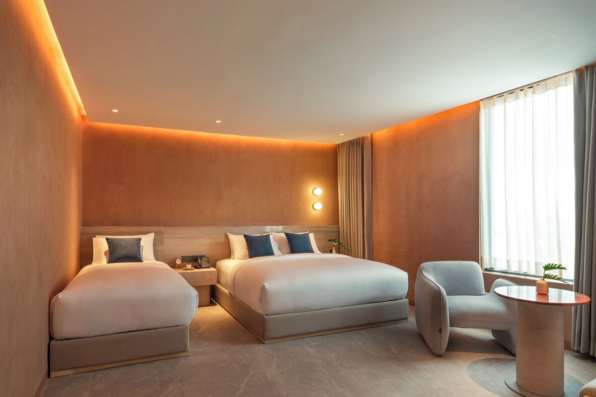 Quarto de hotel com duas camas, área de estar e uma grande janela com cortinas.