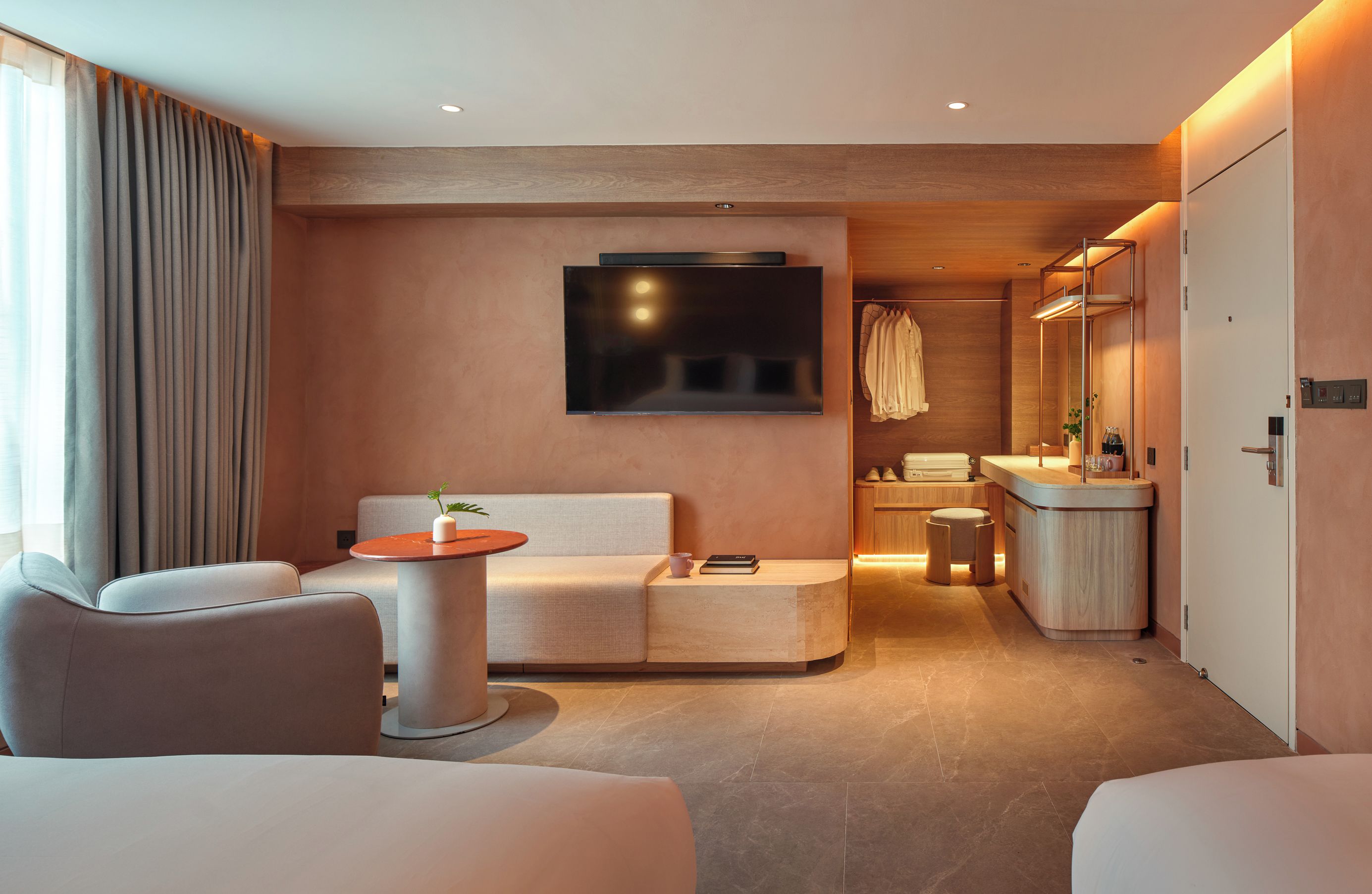 A modern hotel room with a sofa, a small table, a TV, a bathroom with a mirror, and a wardrobe.