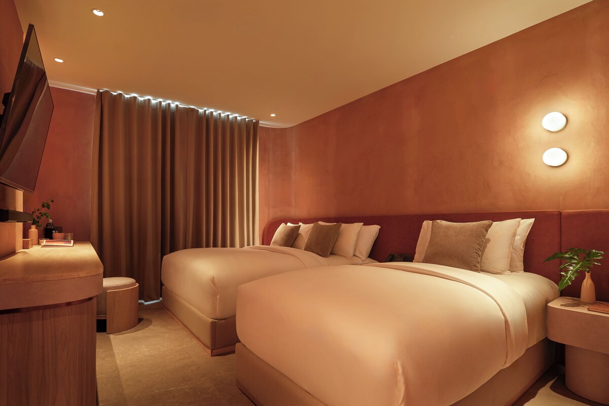 Quarto de hotel com duas camas, televisão, mesa de cabeceira e uma planta.