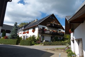 Ein zweistöckiges Gebäude mit Balkon, umgeben von Grün und blauem Himmel.