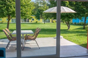 A patio area with a table and chairs, a large umbrella, and a view of a green lawn and buildings in the background. - Bungis holiday home directly on the lake, H1 (Joachimsthal)