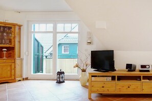 A living room with a television, a small speaker, and a cabinet. There is a large window with a view of a house outside. - Green Street 38 A || Prerow (Prerow)