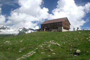 Un rifugio di montagna con facciata in pietra e tetto spiovente, immerso in un pendio erboso con montagne innevate sullo sfondo.