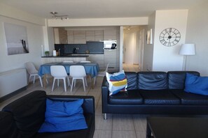 A modern living room with a black leather sofa, a dining area with a table and chairs, and a kitchen in the background. - Holiday apartment with 2 bedrooms (Koksijde)