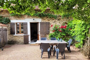 A stone building with a wooden table and chairs outside, surrounded by greenery and flowers. - Authentic gîte with terrace and shared pool (Saint-Saud-Lacoussière)