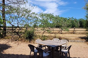 Uma área de refeições ao ar livre com mesa e cadeiras de madeira, cercada por grama seca e árvores.