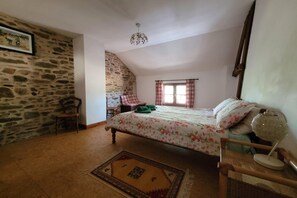 Ein Schlafzimmer mit Steinwand, einem Bett mit floralem Bettzeug, einem Holzstuhl, einem kleinen Tisch und einem Fenster mit Vorhängen.