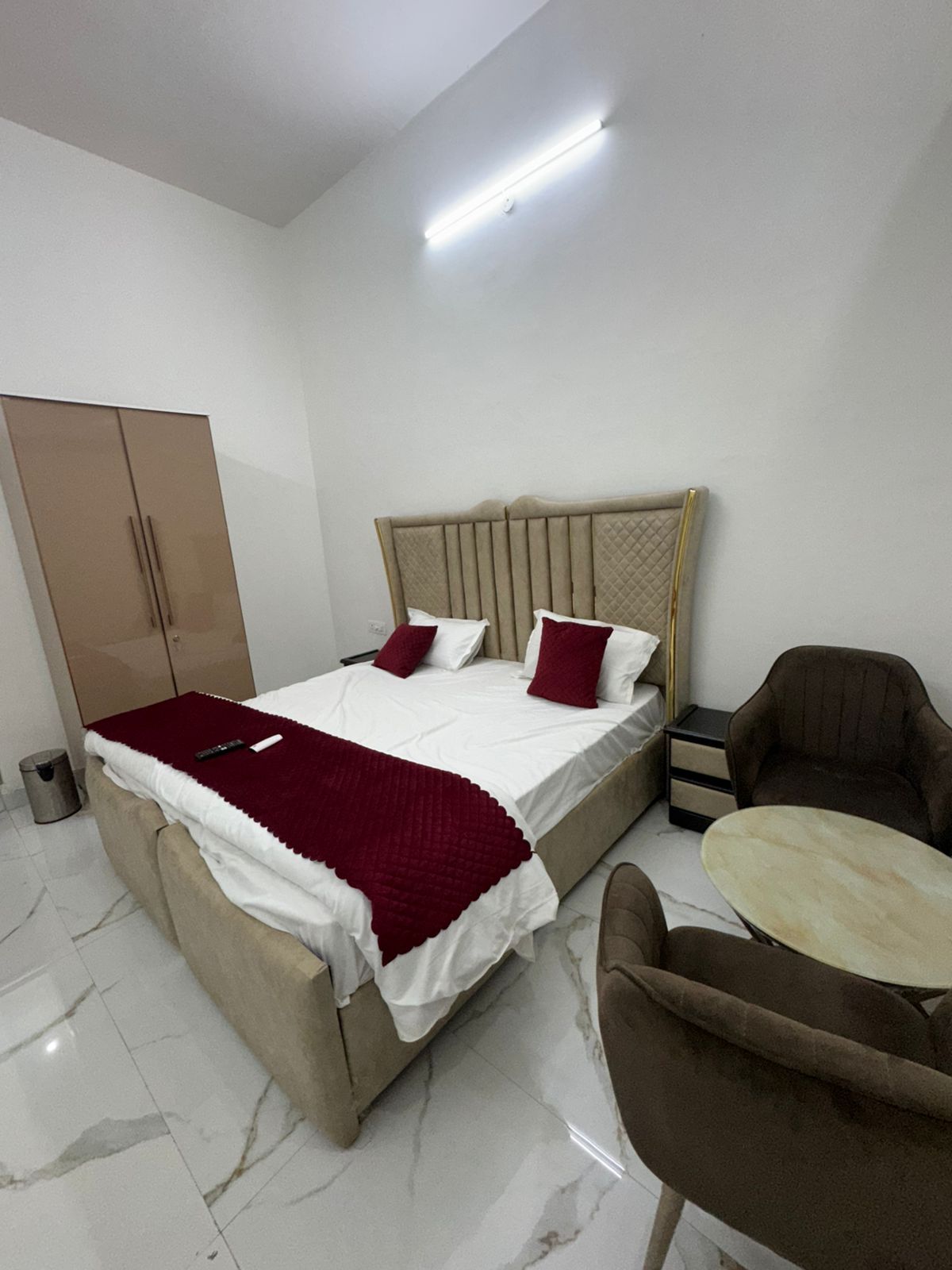 A bedroom with a bed, a wardrobe, a chair, a small table, and a trash bin.