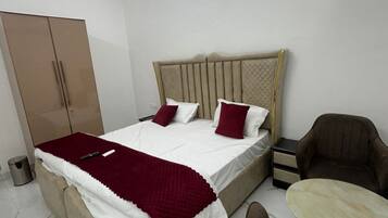 A bedroom with a bed, a wardrobe, a chair, a small table, and a trash bin.