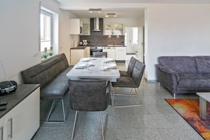 Una sala de estar moderna con un sofá gris, una mesa de comedor lista para cuatro personas y una cocina al fondo.