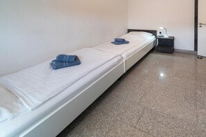 A single-bed room with a white mattress, a small bedside table with a lamp, and a door to the right. - Holiday apartment with 1 bedroom (Bensersiel)
