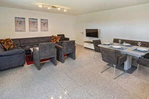 A modern living room with a grey sofa set, a dining table with chairs, a flat-screen TV, and artwork on the wall. - Holiday apartment with 1 bedroom (Bensersiel)