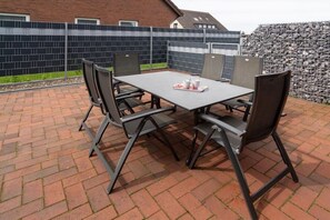 An outdoor patio with a rectangular table and four chairs, a tiled wall, and a stone wall in the background. - Holiday home with 1 bedroom (Neuharlingersiel)