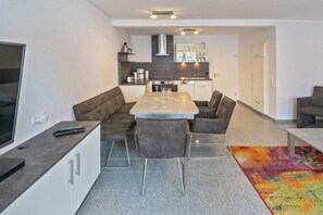 A modern kitchen with a central island, a dining area with a sofa and chairs, and a television on a stand. - Holiday home with 1 bedroom (Neuharlingersiel)