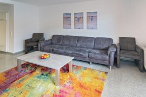 A living room with a gray sofa, two armchairs, a coffee table with fruit, and a colorful rug. - Holiday home with 1 bedroom (Neuharlingersiel)