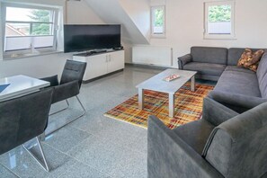 Een moderne woonkamer met een grijze bank, een flatscreen televisie, een eettafel met stoelen en een kamerkleed met een patroon.