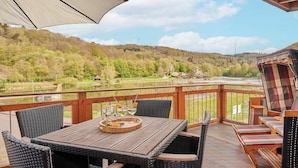 An outdoor deck with a wooden table and wicker chairs, overlooking a lake and forested hills. - Holiday apartment Mila OG (Rieden)