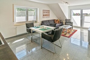 A modern living room with a dining table, chairs, a sofa, and a large window. - Holiday apartment with 1 bedroom (Bensersiel)