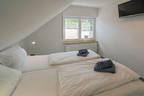 A bedroom with two beds, a window with blinds, a radiator, and a TV mounted on the wall. - Holiday apartment with 1 bedroom (Bensersiel)