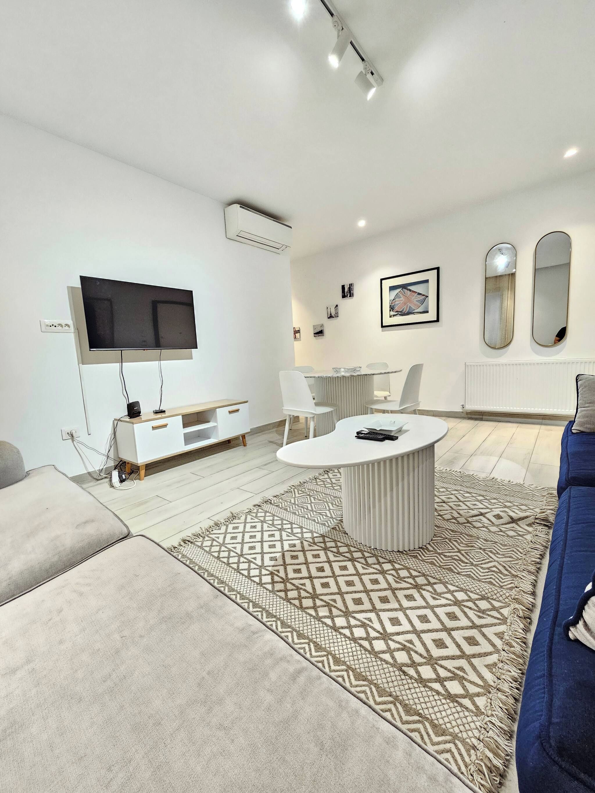 A modern living room with a flat-screen TV, a white entertainment unit, a round coffee table, and a patterned rug.
