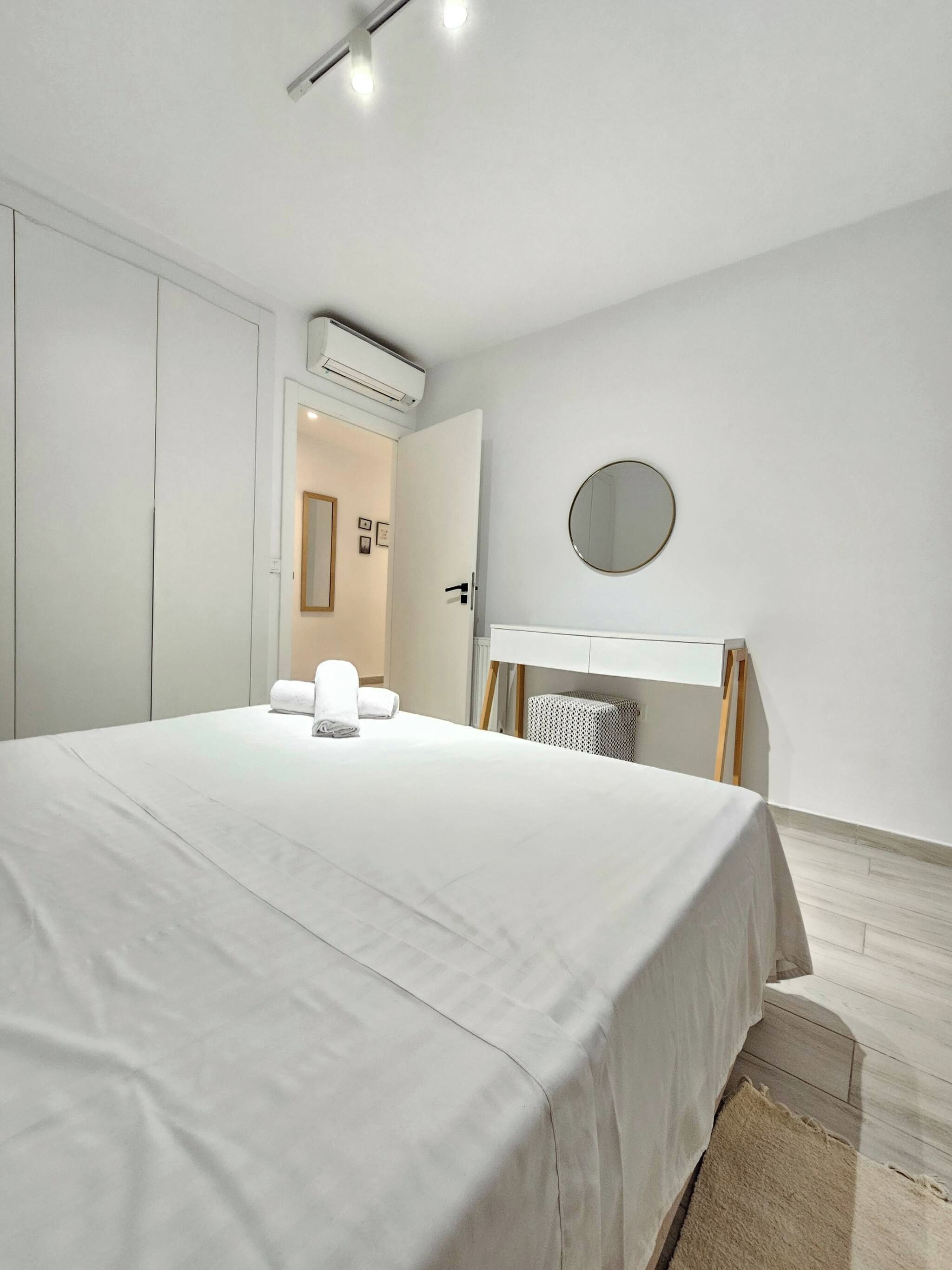 A modern bedroom with a large bed, a bedside table with a mirror, and a white wardrobe.