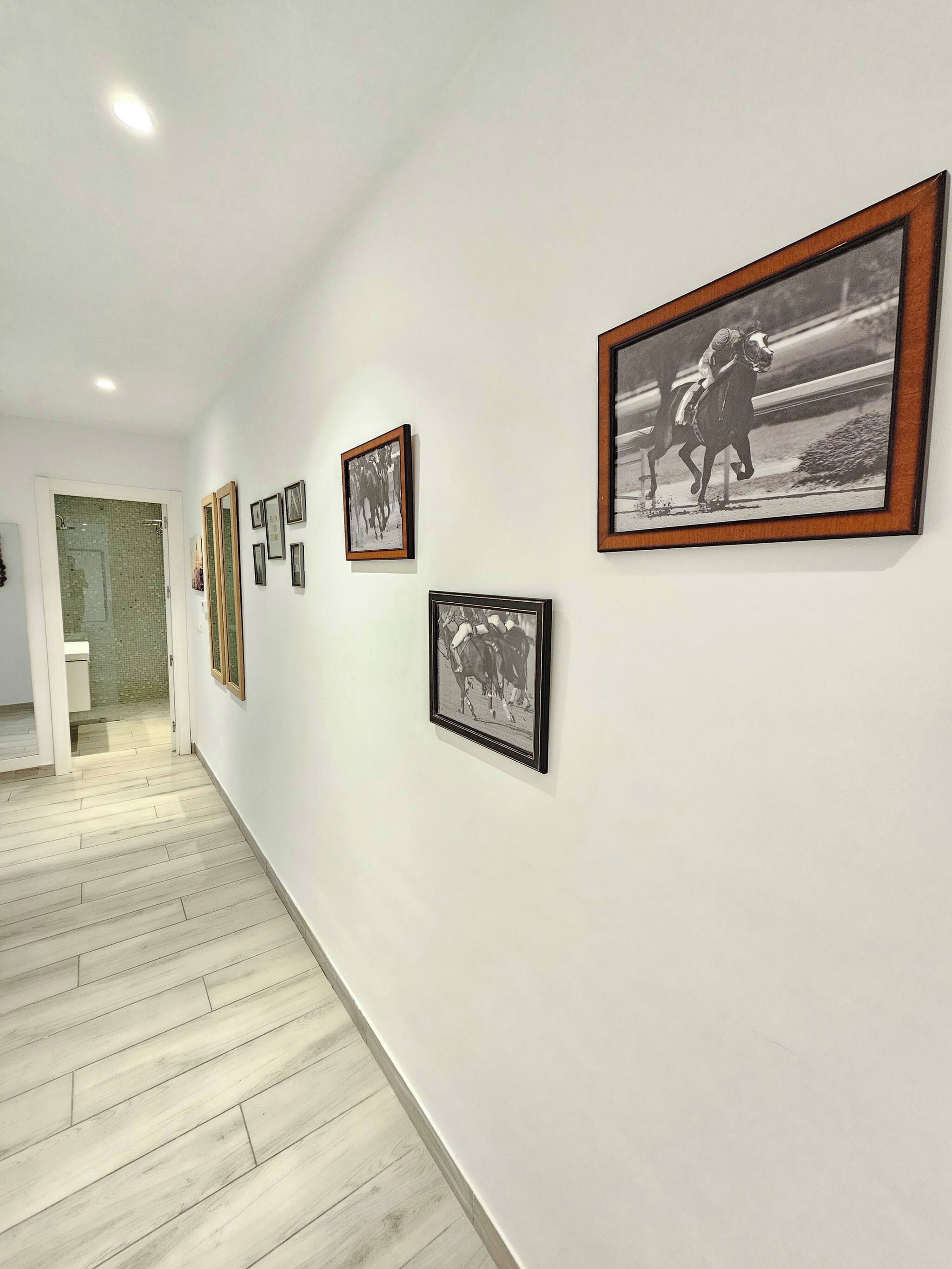 A hallway with framed black and white photographs on the walls, a wooden floor, and a door leading to a bathroom.