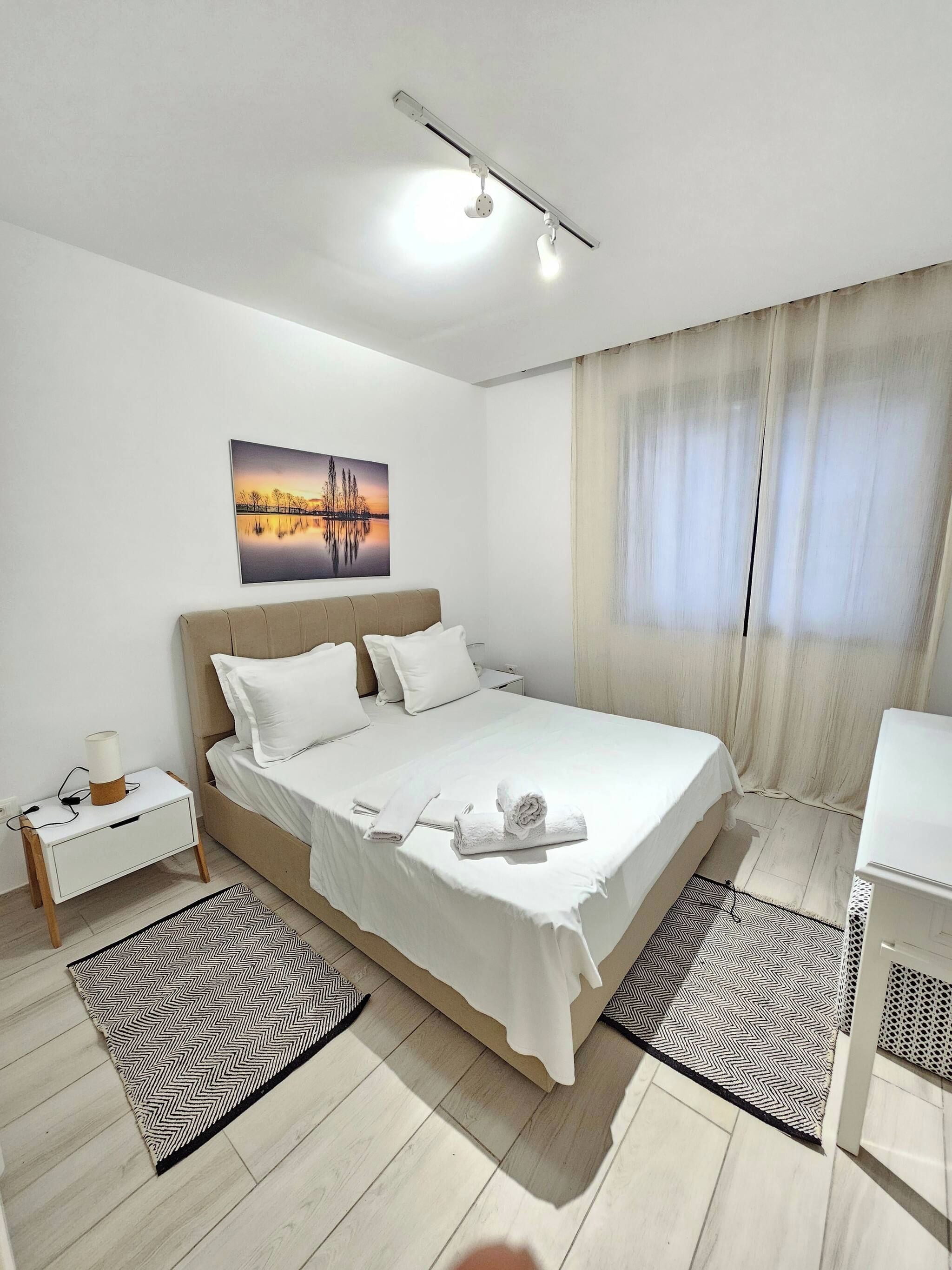 A hotel room with a bed, bedside tables, a nightstand, a painting on the wall, and a window with curtains.