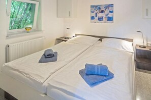 A double bed with white linens, two bedside tables with lamps, a window with a view of trees, and a painting on the wall. - Holiday apartment with 1 bedroom (Bensersiel)