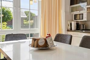 Ein Esstisch mit Kuchen und einer Kaffeetasse in einem Raum mit Fenster, Vorhang und einer Küchenzeile mit Geräten.