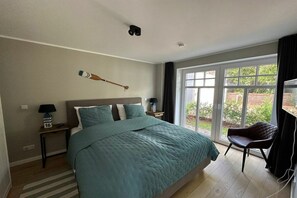 A bedroom with a large bed, a nightstand, a chair, and a window with a view of greenery. - Holiday apartment with 1 bedroom (Wangerooge)