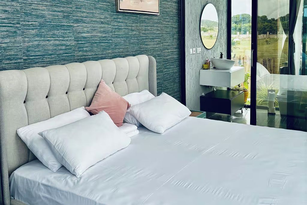 Une chambre à coucher moderne avec une tête de lit capitonnée, du linge de lit blanc et une vue sur un jardin, visible à travers une porte vitrée.