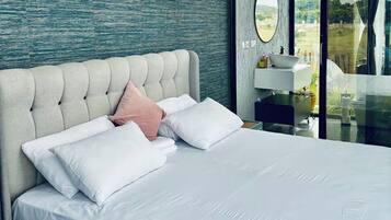 Une chambre à coucher moderne avec une tête de lit capitonnée, du linge de lit blanc et une vue sur un jardin, visible à travers une porte vitrée.