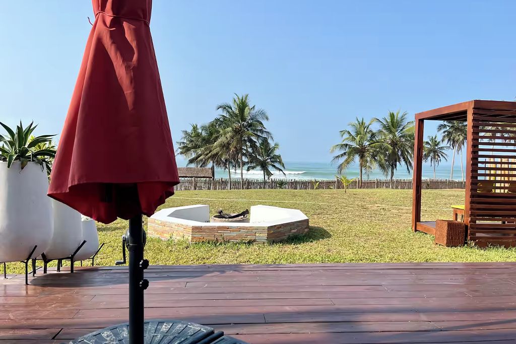 Une terrasse en bois avec un parasol rouge, un foyer et une vue sur la plage, où l’on aperçoit des palmiers.