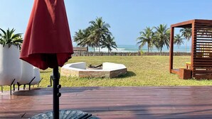 Una terraza de madera con un paraguas rojo, una chimenea y vistas a la playa con palmeras.