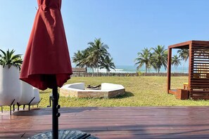 A wooden deck with a red umbrella, a fire pit, and a view of the beach with palm trees.