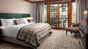 Ein Hotelzimmer mit einem großen Bett, einem Sessel, einem Schreibtisch und Blick auf Gebäude durch eine Glastür.