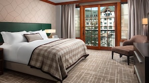 Quarto de hotel com cama grande, uma cadeira, uma escrivaninha e vista para edifícios através de uma porta de vidro.
