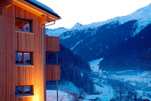 A modern wooden building with multiple windows, set against a snowy mountainous backdrop. - Chalet with fireplace and garden (Gaschurn)