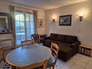 A living room with a brown sofa, a dining table with chairs, a small side table, a bookshelf, and a painting on the wall. - 1-Bedroom Apartment in JARDINS DU GOLFE with Community Pool (Le Plan-de-la-Tour)