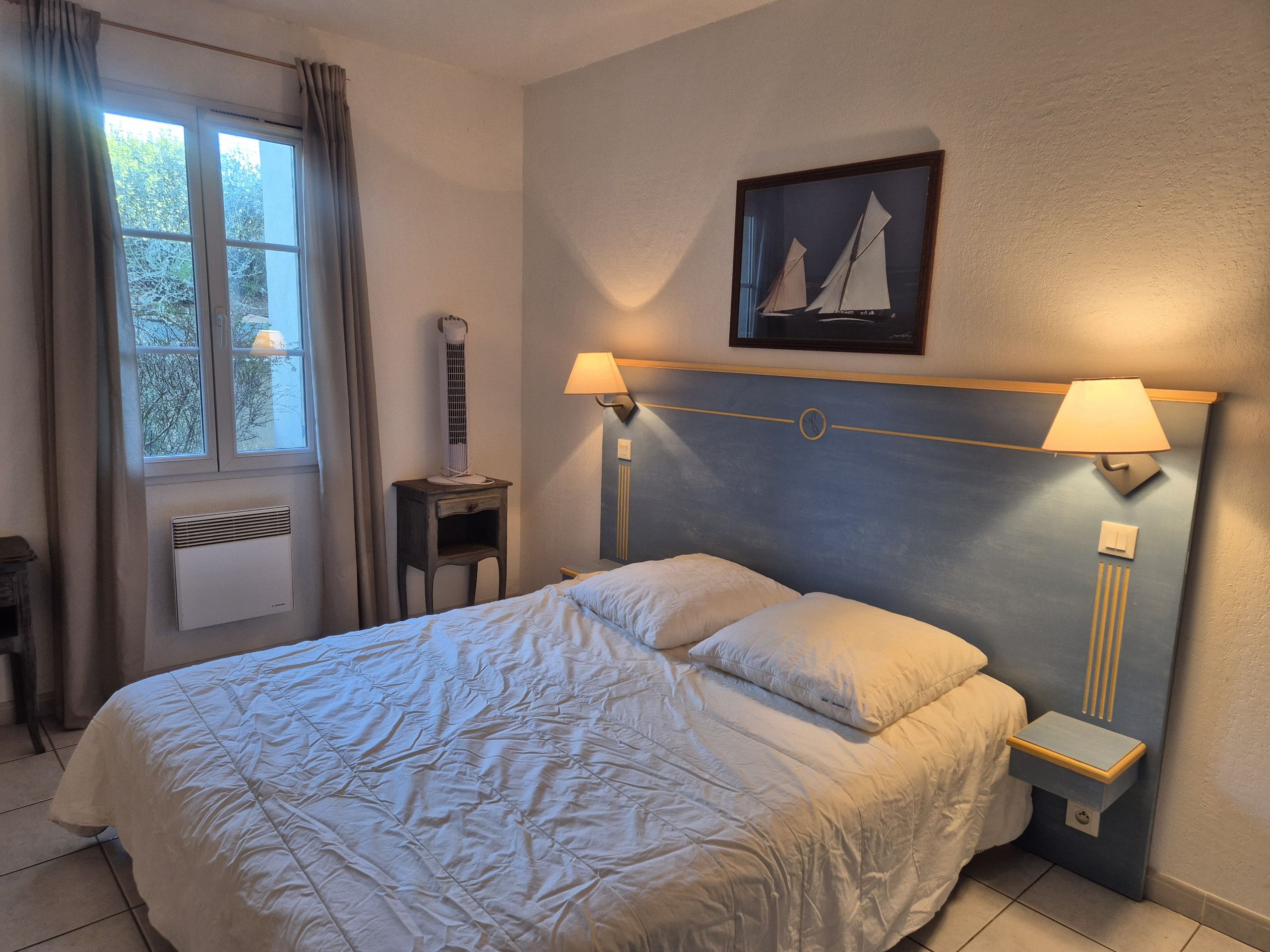 Een hotelkamer met een bed, nachtlampjes, een verwarming, een raam met gordijnen en een schilderij van een zeilboot aan de muur.