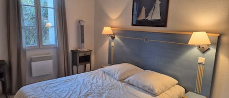 A hotel room with a bed, bedside lamps, a heater, a window with curtains, and a sailboat painting on the wall.