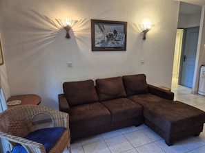 A living room with a brown sectional sofa, a wicker chair with a blue cushion, and two wall-mounted lamps. - 1-Bedroom Apartment in JARDINS DU GOLFE with Community Pool (Le Plan-de-la-Tour)