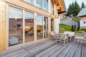 A wooden deck with outdoor furniture, large sliding glass doors leading to an interior space, and a view of a residential area with trees. - Chalet #2 with 3 bedrooms and sauna (Niederwölz)