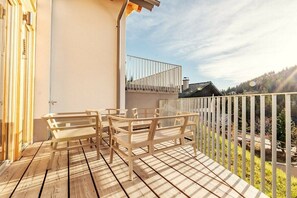 Une terrasse en bois avec des meubles d’extérieur, un bâtiment blanc et une vue sur une colline boisée.