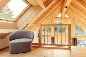 Ein Holzinnenraum mit schrägverdeckter Decke, einem grau gepolsterten Sessel und Blick durch große Fenster.