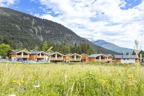 A row of wooden cabins with balconies, set in a meadow with a mountain backdrop.