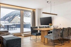 Ein moderner Essbereich mit einem Holztisch und grau gepolsterten Stühlen, einem flachbildfernseher an der Wand und einem großen Fenster mit Blick auf eine Berglandschaft.