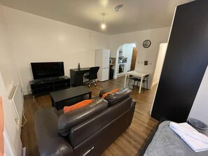 Ein Wohnzimmer mit einer Couch, einem Fernseher, einem Esstisch und einer Küche im Hintergrund.